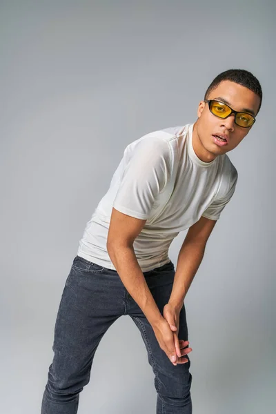 Jeune homme afro-américain en t-shirt élégant, jeans et lunettes de soleil regardant la caméra sur gris — Photo de stock