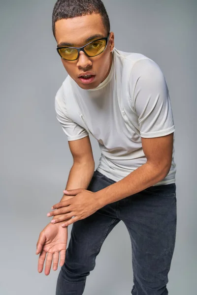 Homme afro-américain confiant en t-shirt à la mode, jeans et lunettes de soleil regardant caméra sur gris — Photo de stock