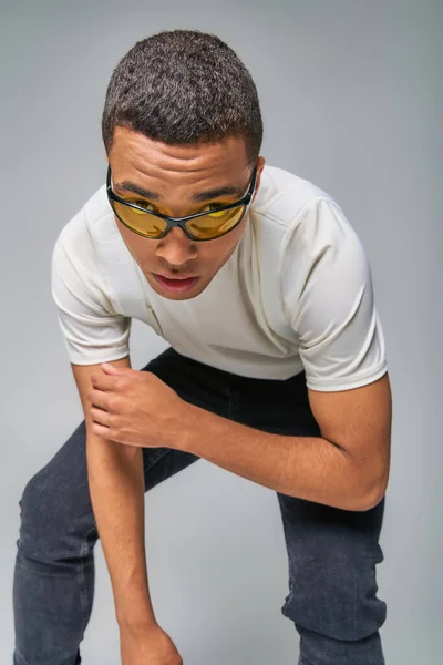 Homme afro-américain expressif en t-shirt à la mode, jeans et lunettes de soleil regardant caméra sur gris — Photo de stock