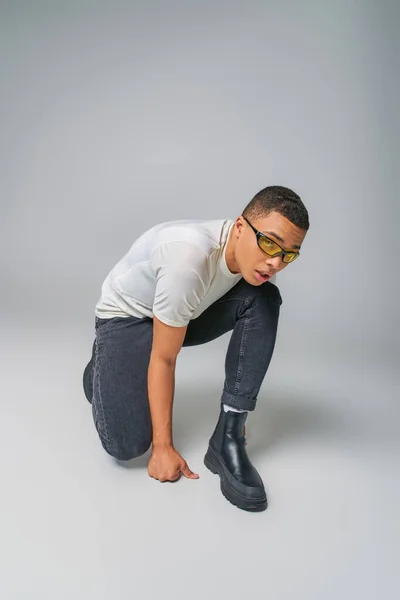 Homme afro-américain avant-gardiste en t-shirt, jeans et lunettes de soleil regardant loin sur gris — Photo de stock