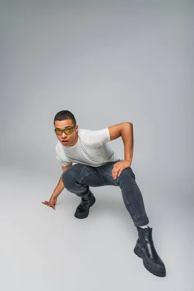 Modern style african american guy in t-shirt, jeans and sunglasses looking at camera on grey — Stock Photo