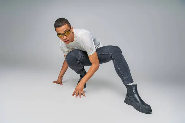 Молодіжна мода, афроамериканський хлопець у футболці, джинсах і сонцезахисних окулярах позує на сірому — стокове фото