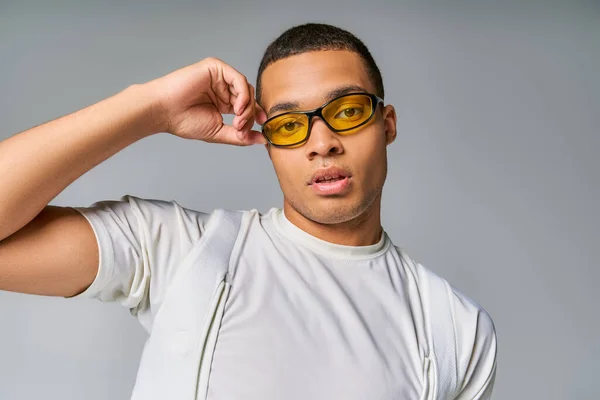 Молодий, стильний афроамериканський чоловік у футболці, що регулює сонцезахисні окуляри, дивлячись на камеру на сірому — стокове фото