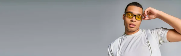 Trendy african american man in t-shirt adjusting sunglasses, looking at camera on grey, banner — Stock Photo