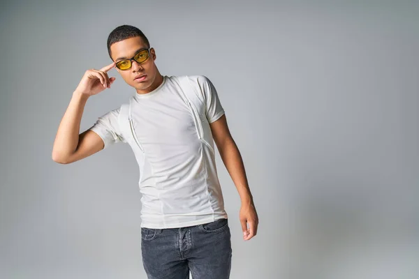 Homem americano africano na moda em t-shirt, jeans e óculos de sol apontando para a cabeça em cinza — Fotografia de Stock