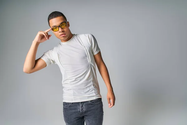 Fashionable african american man in t-shirt, jeans and sunglasses pointing at head on grey — Stock Photo