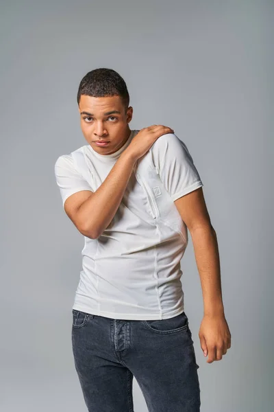 Moda juvenil, expresivo afroamericano en camiseta y jeans mirando a la cámara en gris - foto de stock