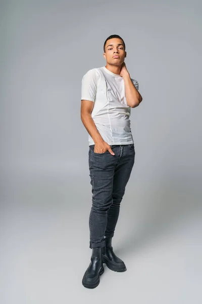 Moderno afroamericano en camiseta y jeans, con la mano en el bolsillo, mirando a la cámara en gris - foto de stock