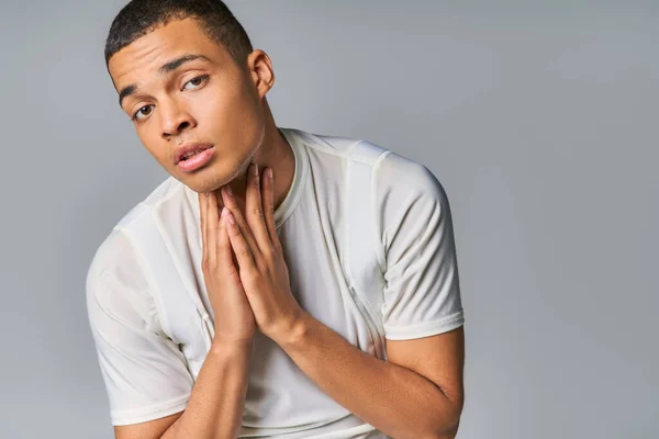 Homme afro-américain à la mode en t-shirt tenant les mains près du cou et regardant la caméra sur gris — Photo de stock