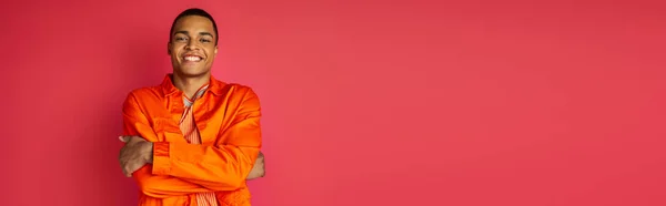 Gioioso uomo afroamericano in camicia arancione, con le mani incrociate, guardando la fotocamera su rosso, banner — Foto stock