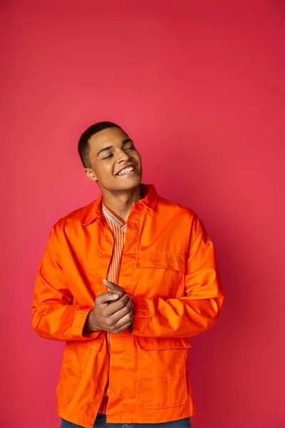 Hombre afroamericano alegre y de moda en camisa naranja mirando hacia otro lado sobre fondo rojo - foto de stock