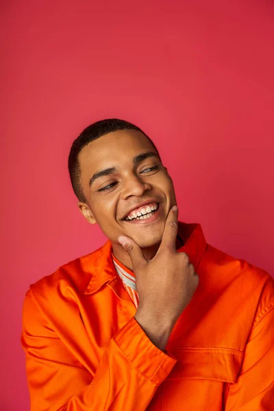 Sonriente y elegante hombre afroamericano en camisa naranja tocando la cara y mirando hacia otro lado en rojo - foto de stock