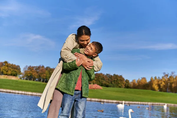 Vínculo entre la madre y el niño, alegre mujer afroamericana abrazando chico, estanque con cisnes - foto de stock