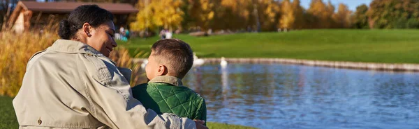 Bonding between mother and child, cheerful african american woman hugging boy, pond, swans, banner — Stock Photo