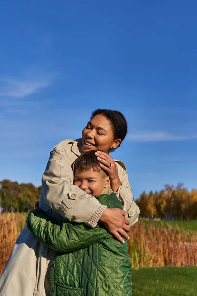 Vinculación, naturaleza otoñal, madre afroamericana feliz abrazando al hijo, familia en ropa de abrigo, caída - foto de stock