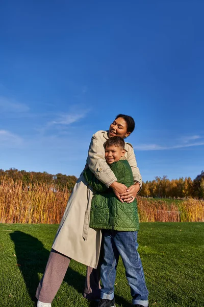 Vinculación, naturaleza otoñal, alegre madre afroamericana abrazando al hijo, familia en ropa de abrigo - foto de stock