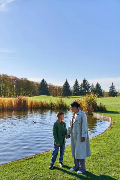 Family bonding, autumnal nature, african american mother walking with son along pond, hold hands — Stock Photo