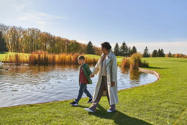 Unión familiar, madre afroamericana caminando con su hijo a lo largo del estanque, cogidas de la mano, otoño, naturaleza - foto de stock