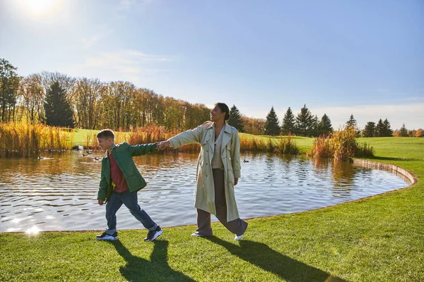 Liens de famille, heureuse femme afro-américaine suivant son fils, marchant le long de l'étang, se tenant la main, automne — Photo de stock