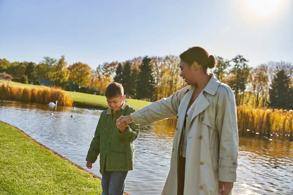 Unión familiar, alegre madre afroamericana caminando con su hijo a lo largo del estanque, cogidas de la mano, otoño - foto de stock