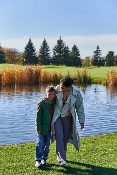 Liens de famille, joyeuse mère afro-américaine marchant avec son fils le long de l'étang, étreinte, saison d'automne — Photo de stock