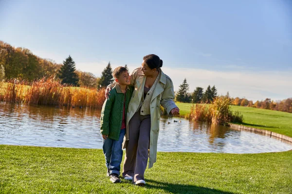Ligação familiar, sorrindo afro-americana mãe andando com o filho ao longo lagoa, abraçando, temporada de outono — Fotografia de Stock