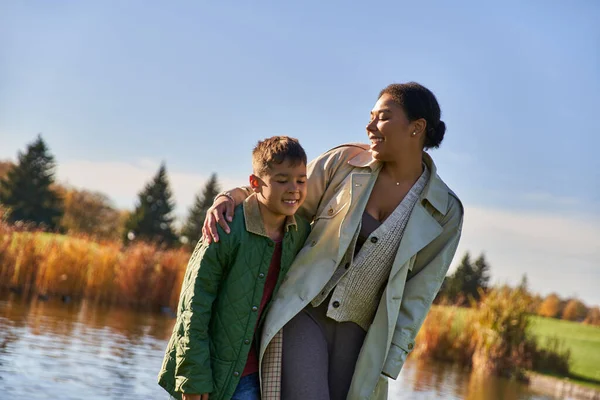Mère afro-américaine positive marchant avec son fils le long de l'étang, étreinte, saison d'automne, famille, liaison — Photo de stock