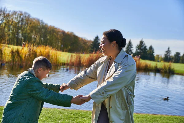 Madre afroamericana feliz cogida de la mano con su hijo cerca del estanque, temporada de otoño, vínculo familiar, risa - foto de stock