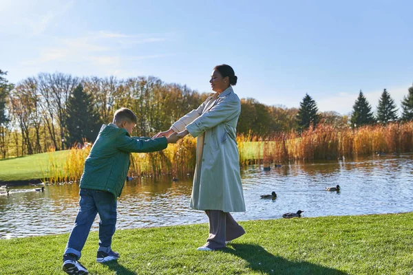 Feliz madre afroamericana cogida de la mano con su hijo cerca del estanque con patos, temporada de otoño, familia - foto de stock
