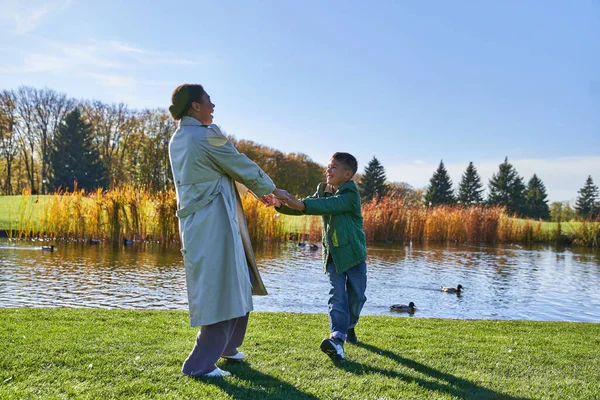 Fröhliche afrikanisch-amerikanische Mutter hält Händchen mit Sohn am Teich mit Enten, verspielt, Herbst — Stockfoto