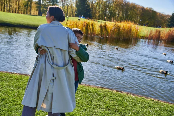 Heureux mère afro-américaine étreignant son fils près de l'étang avec des canards, ludique, saison d'automne, joie, famille — Photo de stock