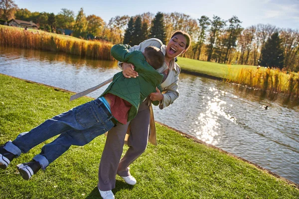 Ludique, joie, heureuse mère afro-américaine levant son fils, s'amuser près de l'étang, vêtements de dessus, automne — Photo de stock
