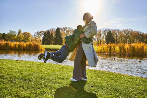 Candid, happy african american mother lifting playful son, having fun near pond, outerwear, autumn — Stock Photo