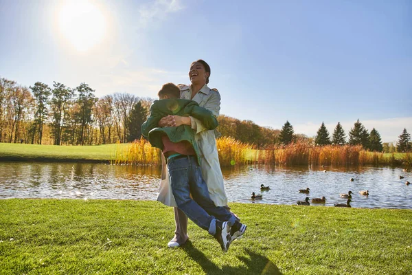 Cándida, temporada de otoño, feliz madre afroamericana levantando a su hijo juguetón, divirtiéndose cerca del estanque - foto de stock