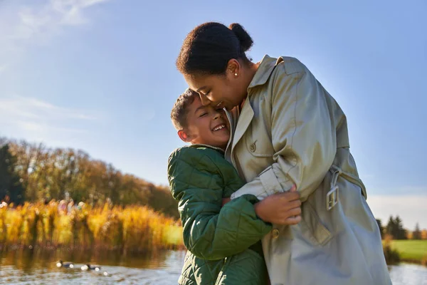 Candid, autumn season, happy african american mother hugging playful son, having fun near pond — Stock Photo