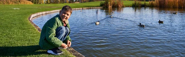 Happy african american boy in outerwear sitting near pond with ducks, nature and kid, banner — Stock Photo
