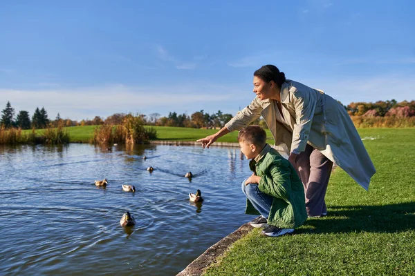 Alegre afroamericana mujer en ropa de abrigo apuntando a patos en estanque cerca de hijo, naturaleza otoñal - foto de stock