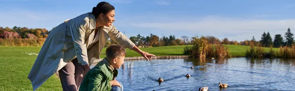 Glücklich afrikanisch-amerikanische Frau zeigt auf Enten im Teich in der Nähe von Sohn, herbstliche Natur, Familie, Banner — Stockfoto