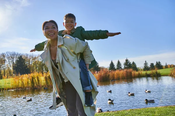 Freudige Mutter huckepack Sohn in der Nähe Teich mit Enten, Kindheit, afrikanisch-amerikanisch, Herbst, frei — Stockfoto