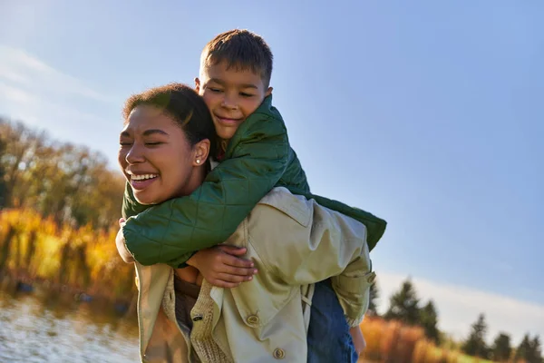 Amor familiar, niño afroamericano alegre abrazando a la madre, naturaleza otoñal, ojos cerrados, felicidad - foto de stock
