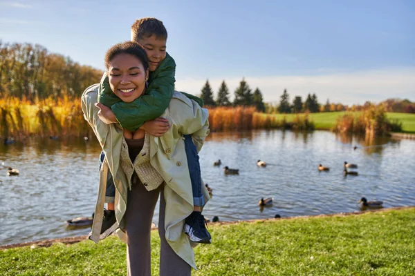 Mãe positiva piggybacking filho perto lagoa com patos, infância, afro-americano, outono, sincero — Fotografia de Stock
