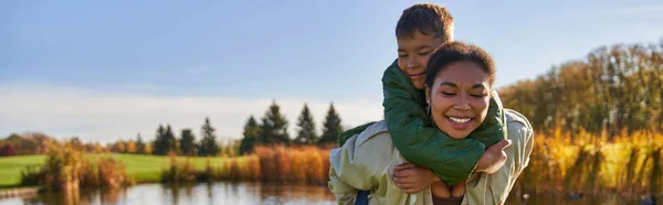 Glückliche Mutter huckepack Sohn am Teich mit Enten, Kindheit, Afroamerikaner, Herbst, Banner — Stockfoto