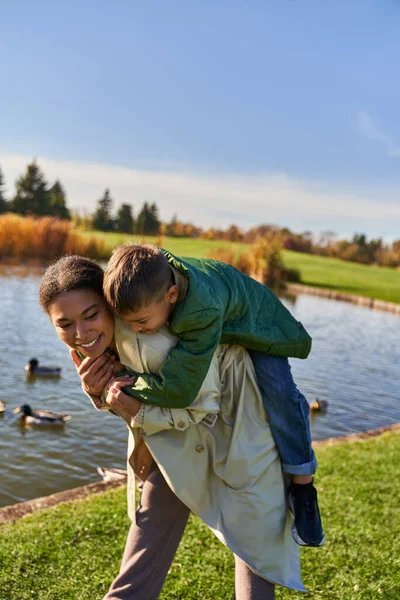 Autunno colori, felice donna africana americana piggybacking figlio vicino stagno con anatre, infanzia, autunno — Stock Photo