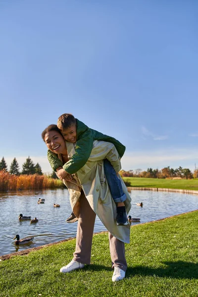 Herbst Farben, fröhliche afrikanisch-amerikanische Frau huckepack Junge in der Nähe Teich mit Enten, Kindheit, Herbst — Stockfoto