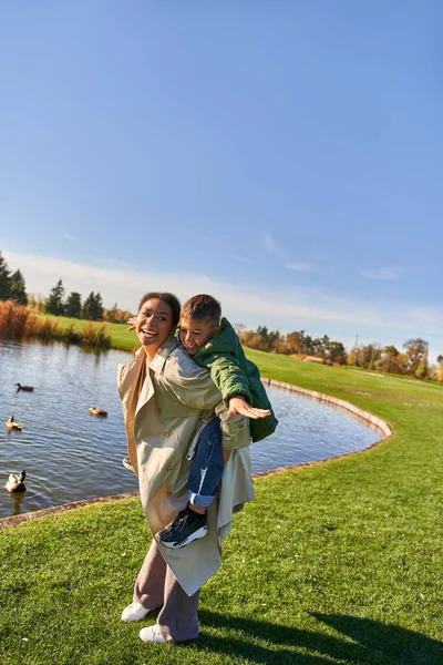 Cores da queda, mãe piggybacking filho perto da lagoa com patos, infância feliz, afro-americana, outono — Fotografia de Stock