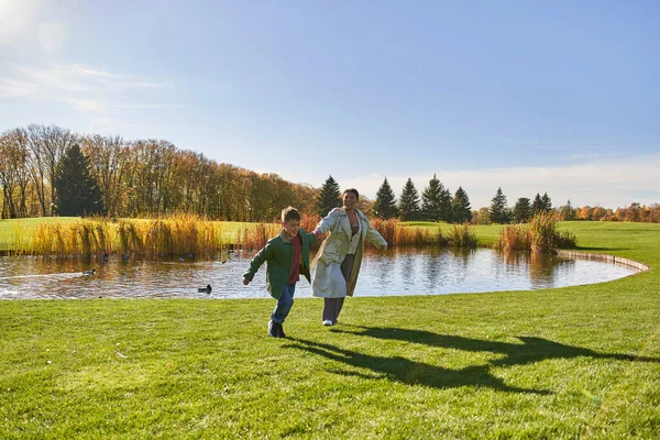 Brincalhão, feliz afro-americano mãe e filho correndo na grama perto da lagoa, parentalidade moderna, diversão — Fotografia de Stock