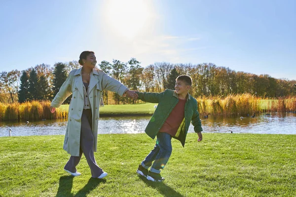 Cheerful african american mother following son, walking near pond, modern parenting, fall fashion — Stock Photo