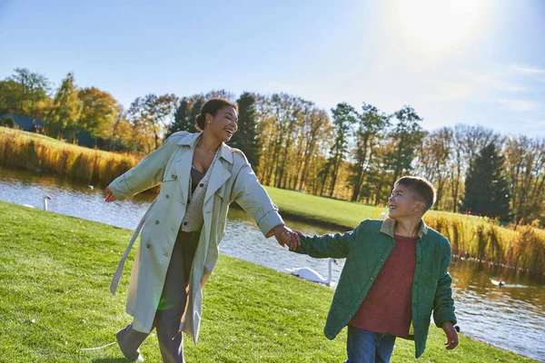 Happiness, african american mother following son, walking near pond, modern parenting, fall fashion — Stock Photo