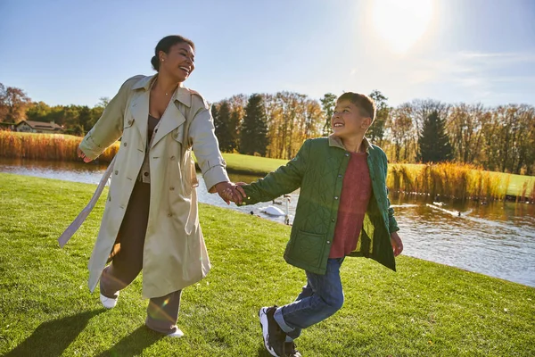 Bonne enfance, femme afro-américaine courant avec son fils près de l'étang, vêtements de dessus, automne, saison d'automne — Photo de stock