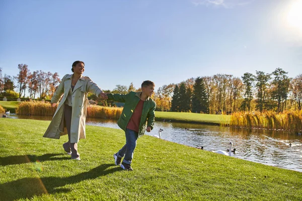 Glückliche Kindheit, Afroamerikanerin in Oberbekleidung läuft mit Sohn in Teichnähe, Herbst, Herbstsaison — Stockfoto